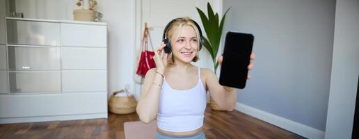 retrato de ajuste y sano mujer rutina de ejercicio a hogar, vistiendo auriculares, demostración teléfono inteligente pantalla, vacío oscuro monitor foto