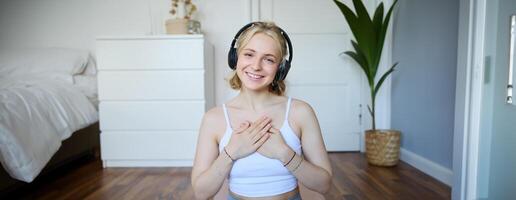 Portrait of woman feeling relaxed and in peace after meditation or yoga training at home, holding hands on chest, wearing headphones, smiling at camera photo