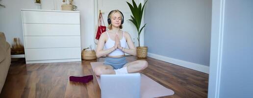 Portrait of young, relaxed woman in headphones, listens to meditation music on laptop, practice yoga on rubber mat, holding hands clasped together in namaste sign photo