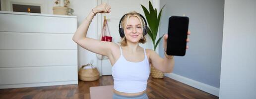 Smiling blond woman working out, showing biceps, recommending smartphone app, demonstrating mobile phone screen at camera, sitting on rubber yoga mat in a room photo