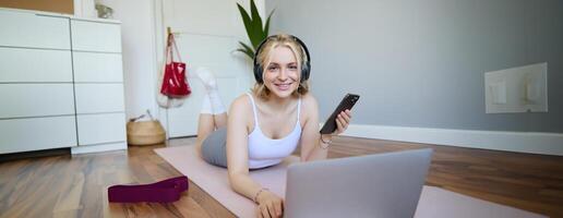 Portrait of happy blond woman in headphones, lying on rubber mat, listens to music in headphones after workout training session, using laptop photo