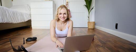 retrato de hermosa rubio mujer mirando a aptitud tutoriales en computadora portátil, acostado en caucho yoga estera, siguiendo rutina de ejercicio instrucciones en línea foto