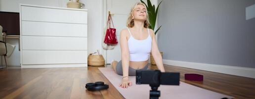 Portrait of young blogger, yoga content creator, showing exercises, recording of herself working out at home on rubber mat photo