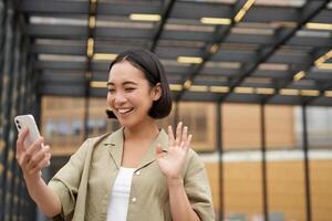 Happy asian girl waves at her smartphone camera, says hello to friend on chat, calling someone, standing on street photo