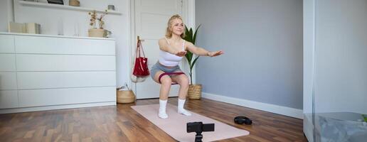 Portrait of young woman, fitness instructor making a for sport vlog, doing squats on camera, using resistance band, working out at home photo