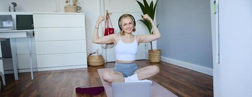 Portrait of young happy woman, fitness blogger in wireless headphones, working out at home, sits on rubber mat with laptop and wireless headphones, shows her muscles photo