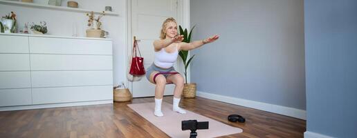 Portrait of woman, fitness instructor at home, recording about workout, showing how to do leg exercises, squats with elastic resistance band, working out indoors on yoga mat photo