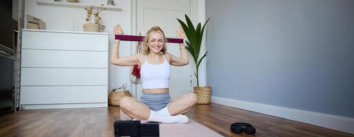 Portrait of young blond woman, social media vlogger using digital camera during workout, shooting about exercises and fitness, stretching with rubber resistance band photo