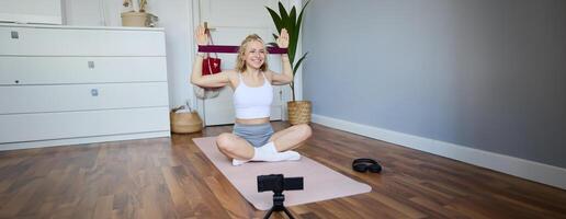 Portrait of young smiling woman, fitness instructor recording about workout, showing how to exercise at home and use rubber resistance band, sitting on yoga mat photo