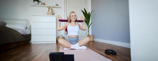Portrait of young blond woman, social media vlogger using digital camera during workout, shooting about exercises and fitness, stretching with rubber resistance band photo