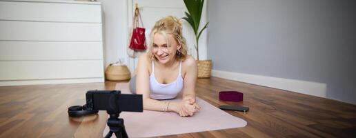 Portrait of young fitness instructor, vlogger creating content at home, doing workout and record exercises on digital camera, using rubber yoga mat photo