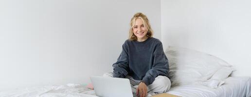 Portrait of young smiling woman studying in her bed, working from home in bedroom, sitting with laptop and notebooks on lotus pose, looking happy and relaxed photo