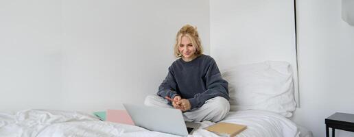 retrato de contento rubio mujer, persona de libre dedicación trabajando desde hogar, sentado en cama con ordenador portátil y cuadernos estudiante haciendo deberes en dormitorio, conecta a en línea clase vía charla foto