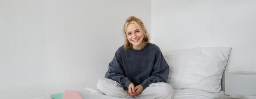 retrato de joven hermosa mujer sentado en cama con ordenador portátil y cuadernos, trabajando desde hogar, trabajo independiente hembra estudiante estudiando en línea, charlas, conecta a lección remotamente foto