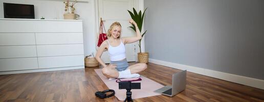 Young female athlete, fitness instructor woman sits on floor rubber mat, recording on digital camera, showing how to workout, explaining exercises photo