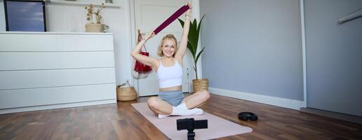 Image of smiling, athletic fitness instructor, female social media influencer, recording about workout, showing how to use resistance bands on digital camera, sitting on rubber yoga mat at home photo