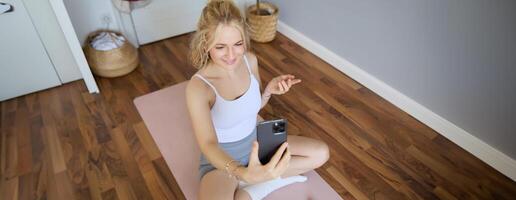 Portrait of young blogger, fitness instructor showing how to do exercises, sitting on yoga mat with smartphone, talking to followers on live stream photo