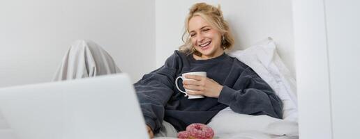 retrato de sincero, contento joven mujer acostado en cama, mirando a ordenador portátil pantalla, participación taza de té y comiendo rosquilla, quedarse a hogar en fin de semana, gasto bastante hora solo foto