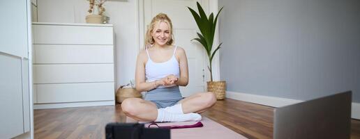 Portrait of young charismatic fitness trainer, girl blogger records on digital camera, talks about health and workout, doing exercises on rubber mat in a room at home photo