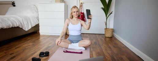 Portrait of beautiful lifestyle blogger, recording herself working out at home, sitting on rubber mat doing yoga in her room, taking selfie on smartphone, using digital camera photo