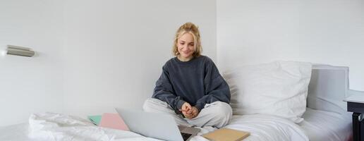 Portrait of young female student, woman studying online, e-learning on her laptop, sitting on bed with notebook and looking at screen, chats, connects to a self-paced course photo