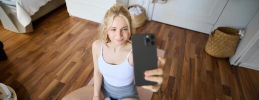 Young sporty fitness trainer, yoga instructor does live stream, takes selfies on smartphone while sitting on rubber mat at home, recording her workout training session photo