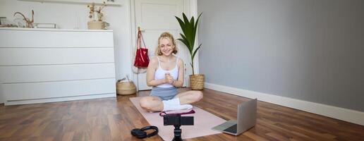 Portrait of young fitness blogger, woman showing exercises to her followers, recording on digital camera, doing workout training session, home yoga, sitting in front of laptop on rubber mat photo