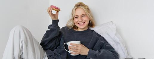 Close up portrait of beautiful smiling, blond woman, lying in bed, drinks tea and eats doughnut on a lazy weekend or day-off photo