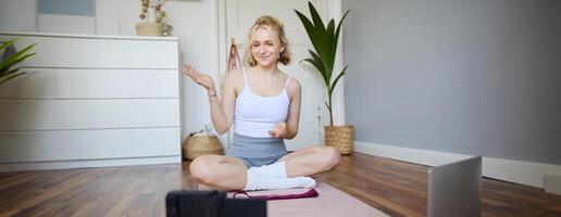 Portrait of beautiful fitness blogger, recording on digital camera, showing workout exercises, explaining fitness movements or yoga training to followers, sits on rubber mat photo