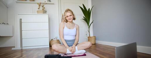 Portrait of young fitness instructor, vlogger showing exercises on camera, recording herself, sitting on mat with laptop, doing workout, explaining yoga movements to followers photo