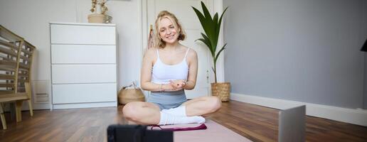 Young female athlete, fitness instructor woman sits on floor rubber mat, recording on digital camera, showing how to workout, explaining exercises photo