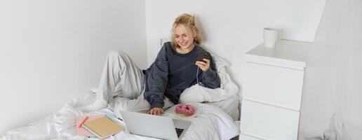 Lifestyle and people concept. Young beautiful woman, staying at home, lying in bed with laptop and smartphone, eating doughnut, enjoying free time, spending weekend at home, watching movie online photo