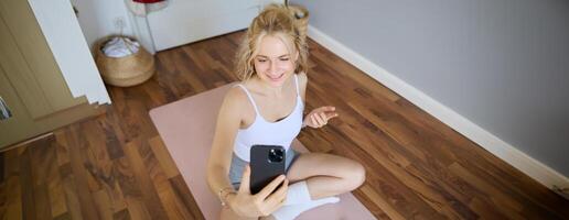 Young sporty fitness trainer, yoga instructor does live stream, takes selfies on smartphone while sitting on rubber mat at home, recording her workout training session photo