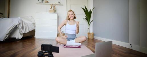 Young female athlete, fitness instructor woman sits on floor rubber mat, recording on digital camera, showing how to workout, explaining exercises photo