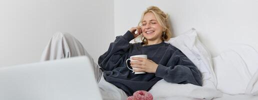 retrato de contento mujer disfrutando día libre a hogar, acostado en cama, acecho televisión espectáculo en computadora portátil, mirando a pantalla y sonriente, Bebiendo té y comiendo en dormitorio foto