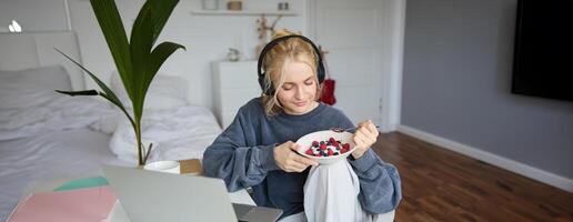 Portrait of smiling young blond woman in headphones, sitting in room, watching movie on laptop, eating breakfast and drinking tea, having lunch in front of computer screen photo