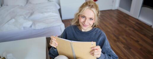 retrato de sonriente, carismático joven mujer, escritura abajo notas, haciendo planes y poniendo eso en planificador, participación diario, sentado en dormitorio y mirando contento a cámara foto