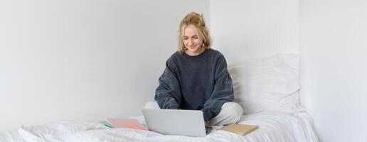 imagen de contento joven mujer, estudiante e-learning desde hogar, conectar a en línea curso en su computadora portátil, se sienta en cama con cuadernos, sonriente y mirando contento foto