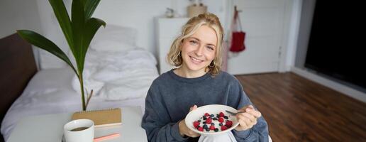 Cute girl eats breakfast and drinks tea in her room, makes herself healthy lunch in bowl, sits in bedroom photo