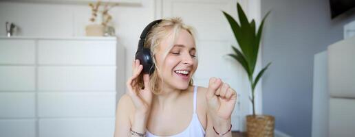 Portrait of happy woman, listens to music in wireless headphones, holding invisible microphone, singing into mic, smiling, enjoying good sound quality photo