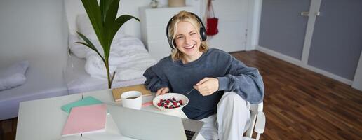 Image of happy woman sitting in a room, watching interesting tv show or movie on laptop, using screaming service, wearing headphones, eating dessert and drinking tea photo