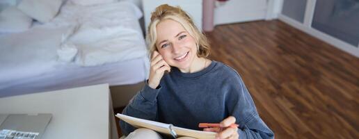 Portrait of smiling, beautiful young candid woman, sitting in bedroom with pen and notebook, writing in journal, making notes, looking happy at camera photo
