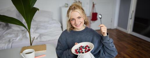 retrato de sonriente rubio mujer, comiendo desayuno, participación cuenco y cuchara, sentado en dormitorio, mirando contento a cámara foto