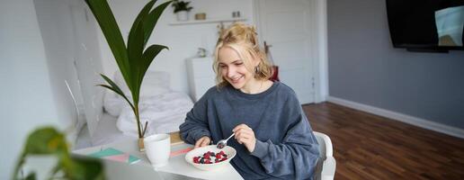 retrato de sonriente rubio joven mujer, comiendo en frente de computadora portátil, acecho s en línea mientras teniendo desayuno, disfrutando postre, sentado en dormitorio foto