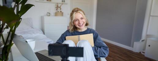 Portrait of cute, smiling young social media content creator, girl records on digital camera and stabiliser, holds notebook, talks to audience, vlogging in her room photo