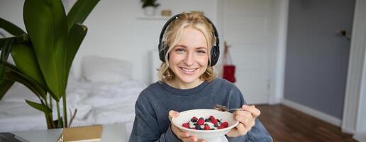 Portrait of smiling cute woman in headphones, eating her breakfast and watching on laptop, looking at screen with happy face photo