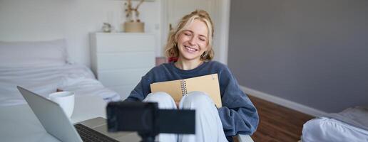 Portrait of blond smiling woman, records on digital camera how she writes in notebook, talks to followers, doing lifestyle blog content in her room photo