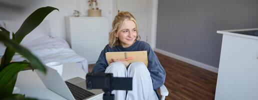 Social media and lifestyle content concept. Young happy smiling woman, sits in her room with notebook, talks at digital camera, having conversation with followers, does lifestyle vlog photo