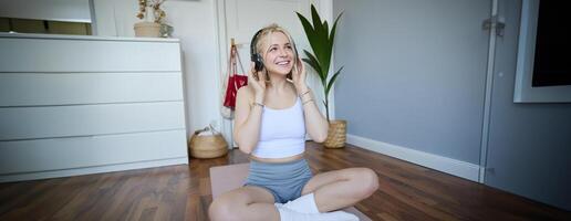 Sport and wellbeing concept. Smiling woman in wireless headphones, sitting on yoga mat, listens to music and workout at home, enjoys fitness training session photo