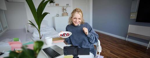 Portrait of smiling, candid young woman, content creator, eating bowl of dessert and looking at digital camera, recording vlog for followers on social media photo